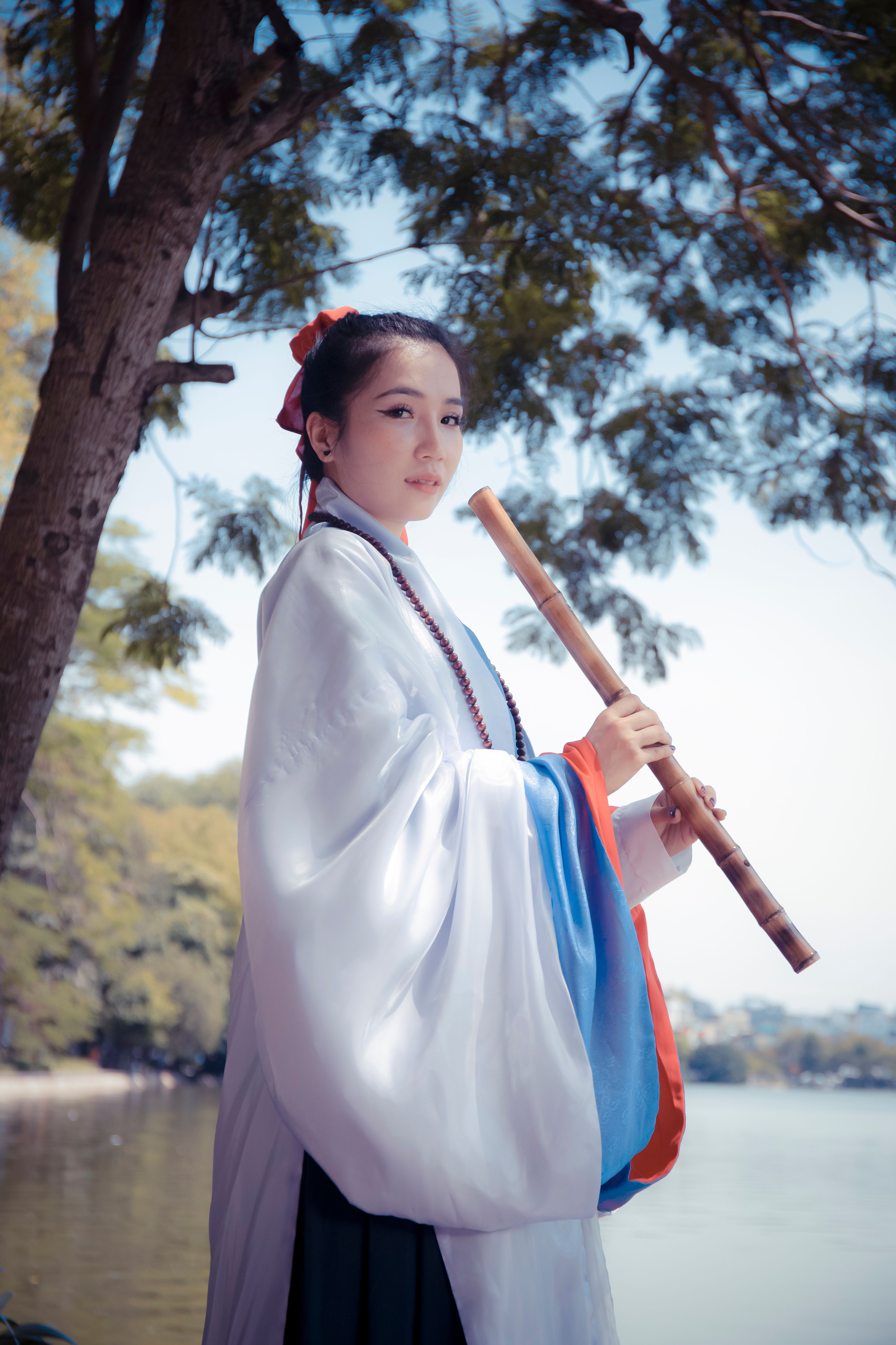 A woman in traditional attire plays a flute by a serene lakeshore, capturing a peaceful moment.