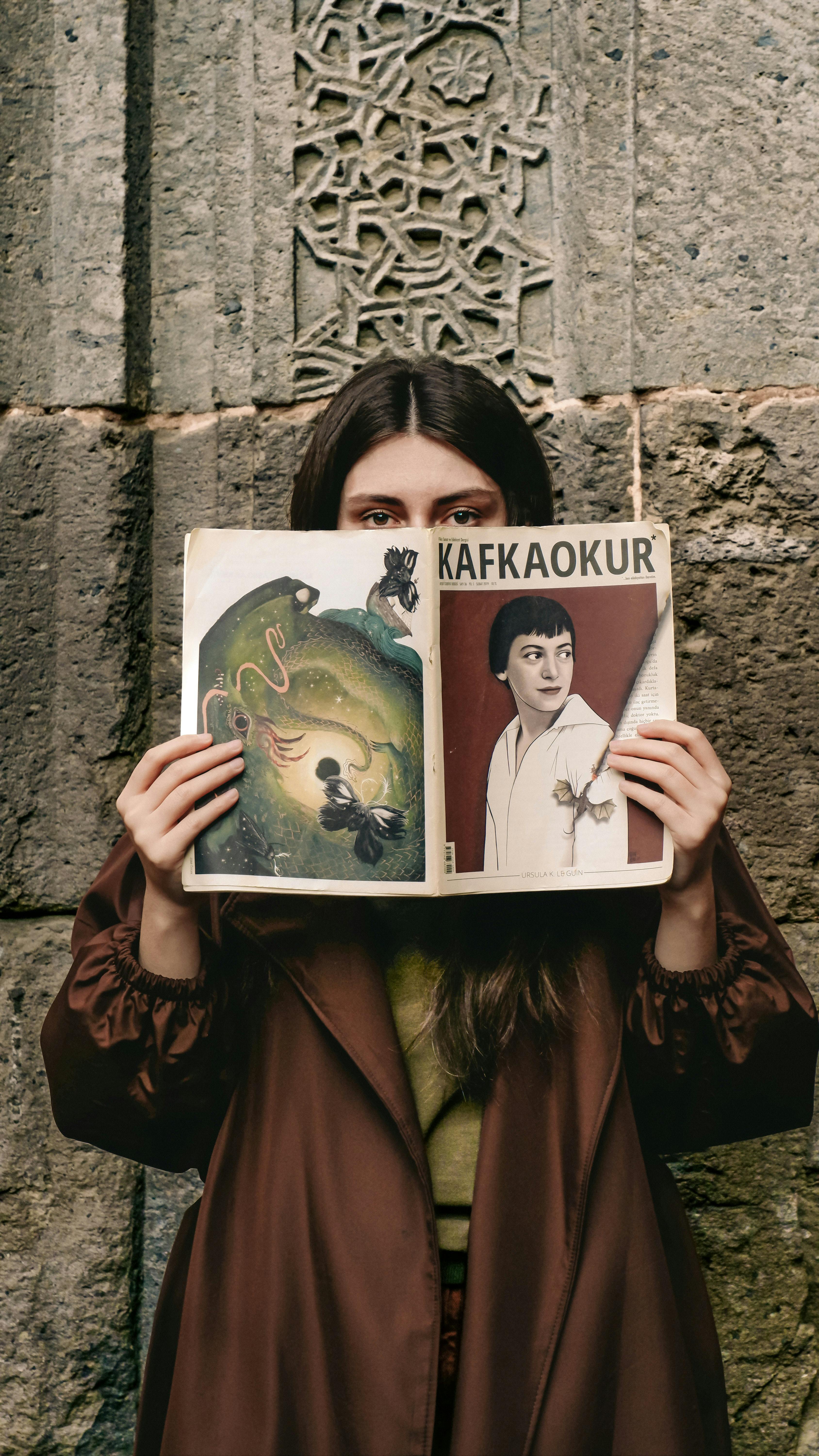 Woman in brown coat holds Kafkaokur magazine against a decorative stone wall.