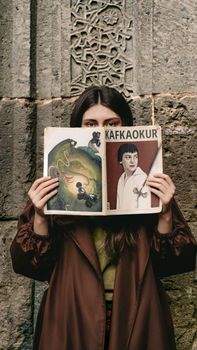 Woman in brown coat holds Kafkaokur magazine against a decorative stone wall.