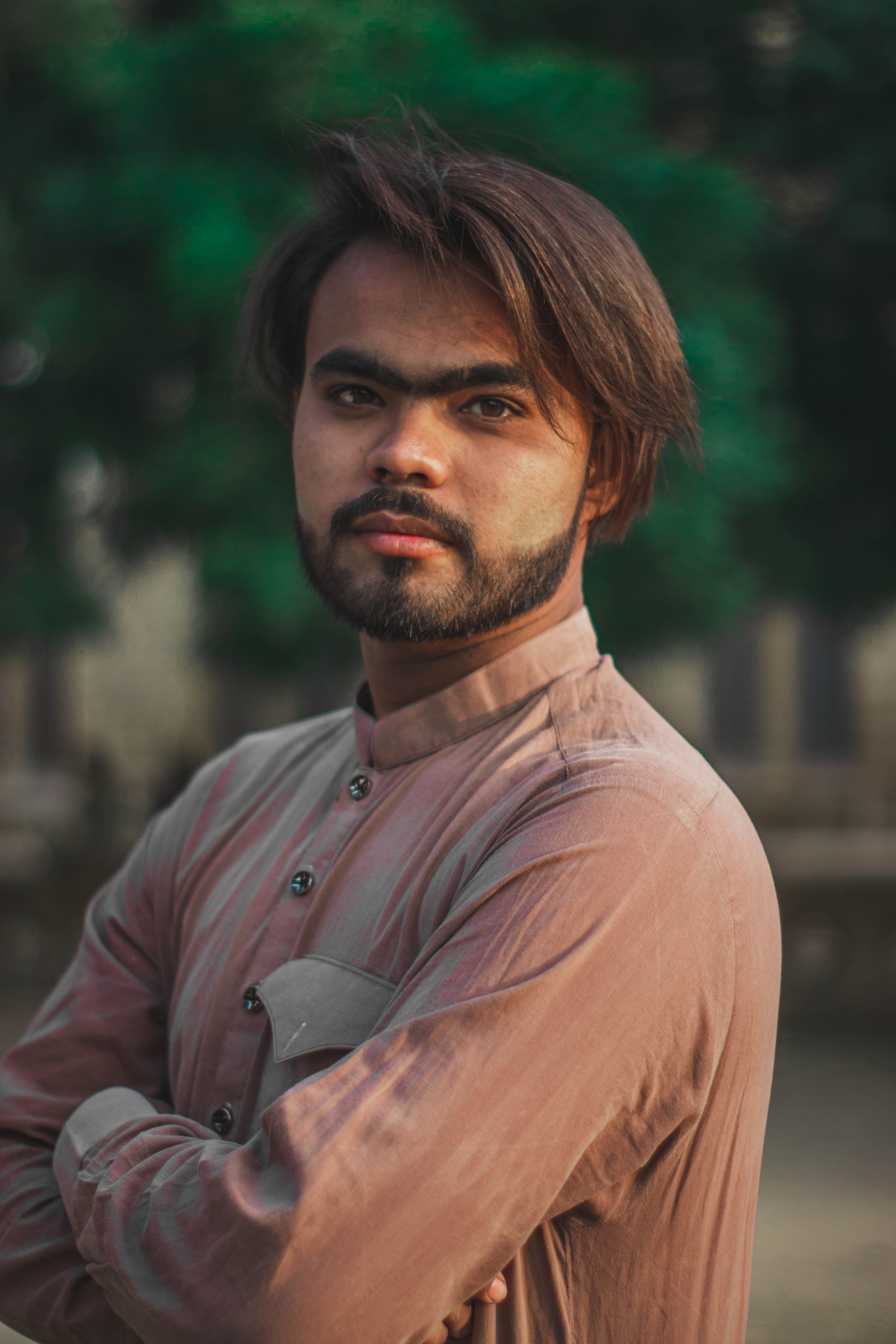 A confident bearded man with a mustache in an outdoor setting, wearing a neutral shirt.