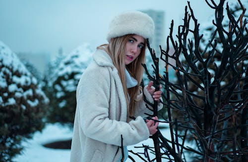 A Woman in White Fur Hat Looking with a Serious Face while Holding Tree Branches