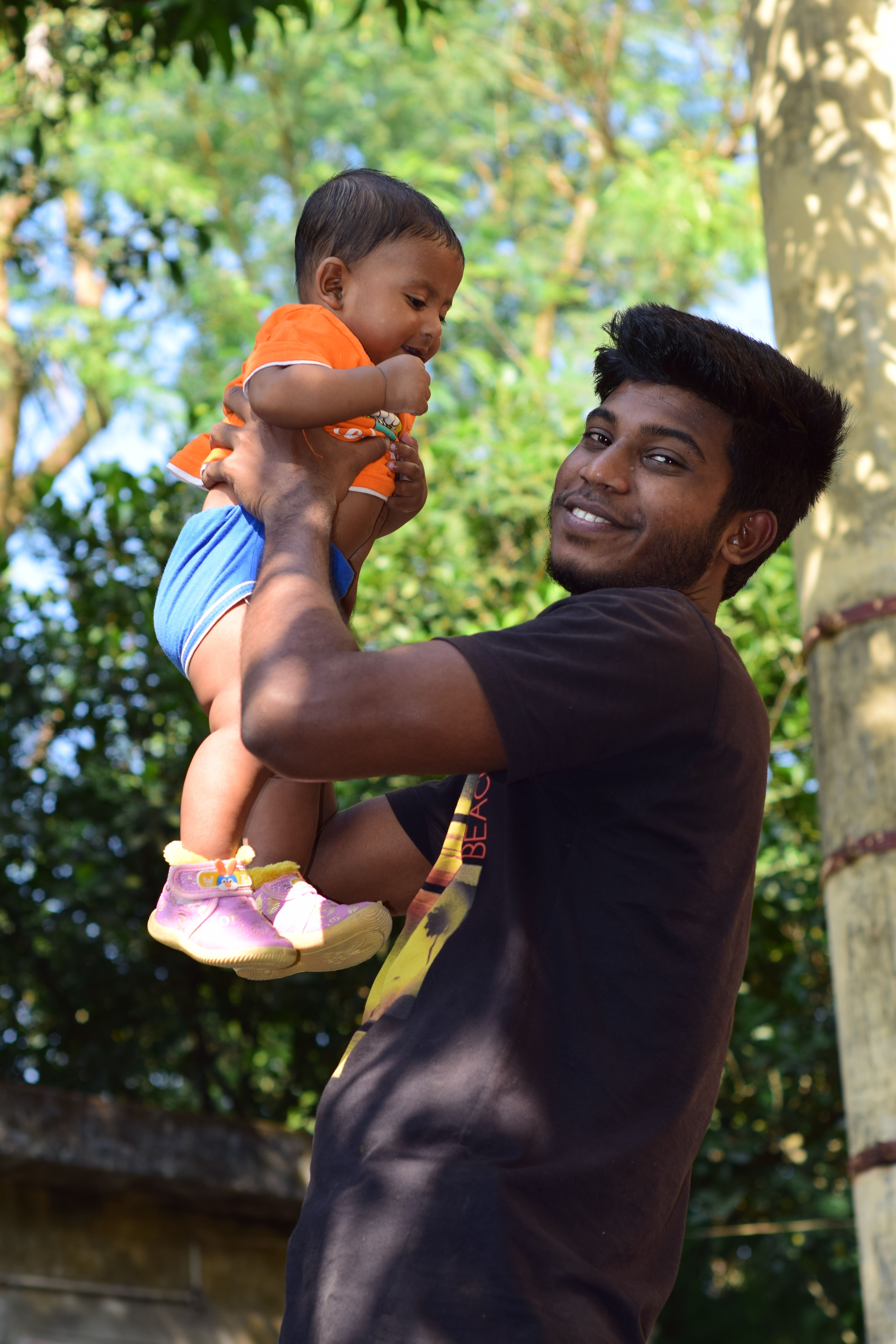 Man joyfully lifting a baby outdoors, surrounded by lush green foliage on a sunny day.