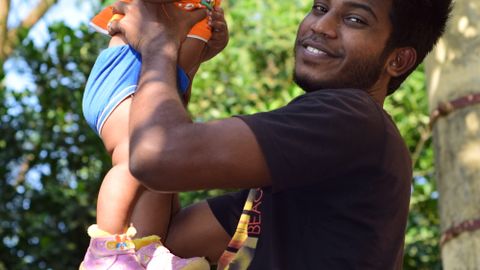 Man joyfully lifting a baby outdoors, surrounded by lush green foliage on a sunny day.