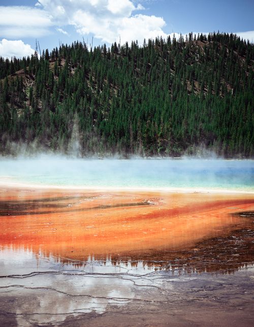 Free Colorful geothermal hot spring surrounded by a forested mountain landscape, emitting steam under a clear sky. Stock Photo