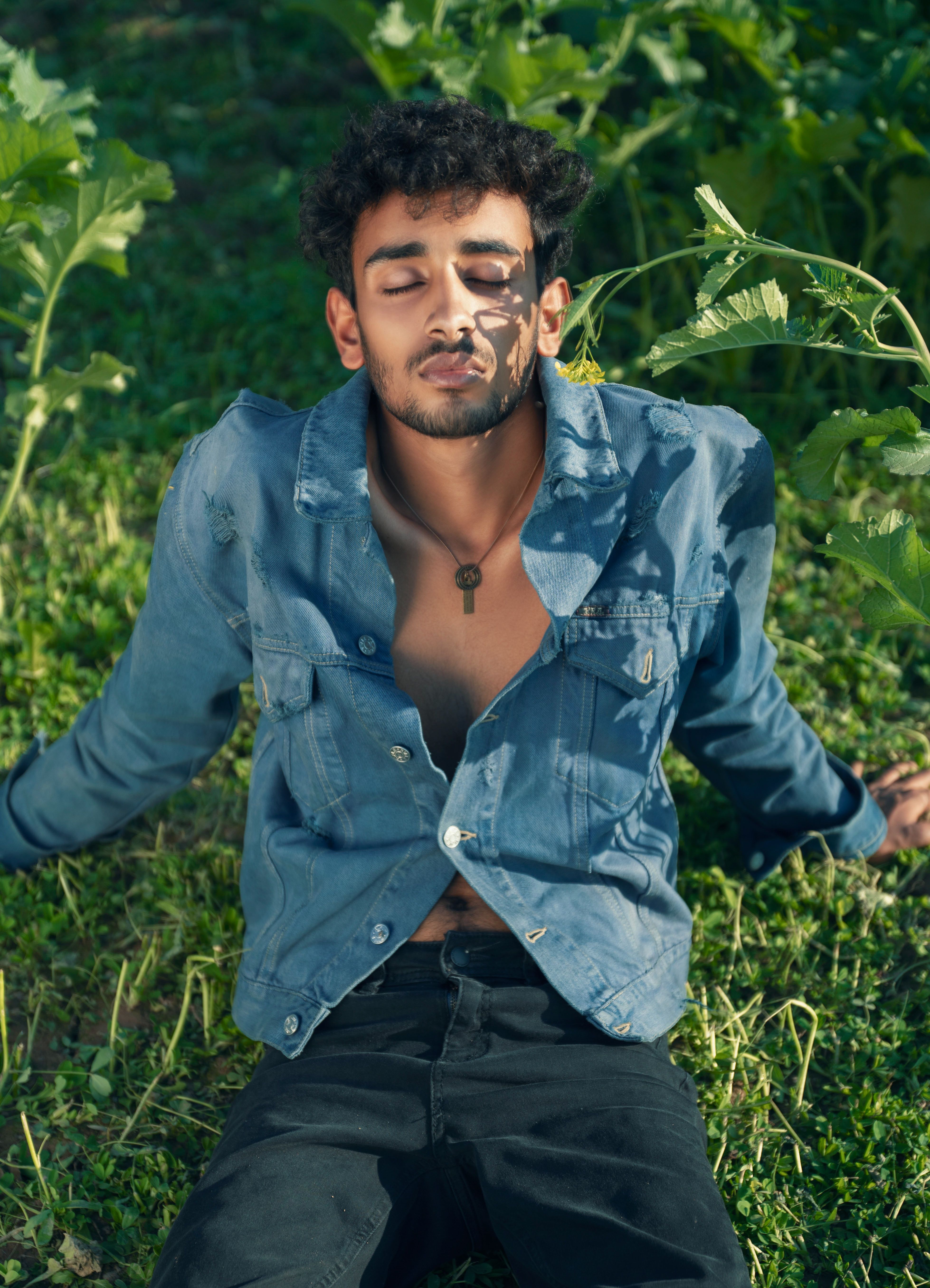 Young man enjoying nature, wearing a denim jacket and black jeans, sitting on grass with relaxed expression.
