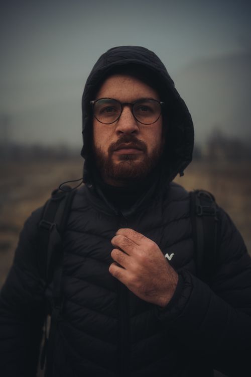 Free A man with glasses and a hooded jacket poses outdoors in a moody setting, capturing an adventurous vibe. Stock Photo