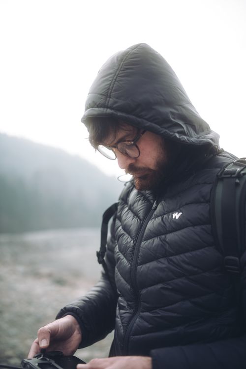 Free Caucasian man wearing glasses and a hooded jacket looking down in misty outdoor setting, suggesting exploration. Stock Photo