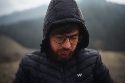 Free Close-up portrait of a bearded man wearing glasses and a hooded jacket in an outdoor setting. Stock Photo