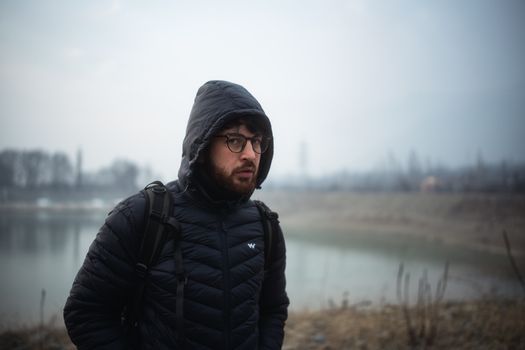 Man wearing glasses and winter jacket standing by a foggy lake, exuding a contemplative mood.