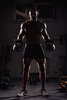 Shirtless man lifting kettlebells in dark gym, showcasing strength and fitness.