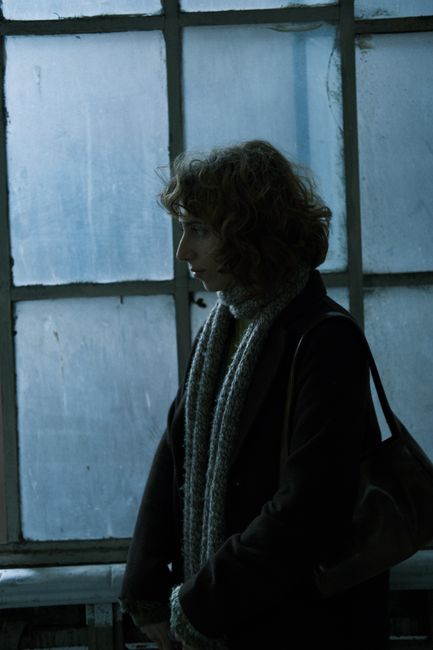 Side profile of a woman with curly hair standing by a frosted window, wearing a scarf.
