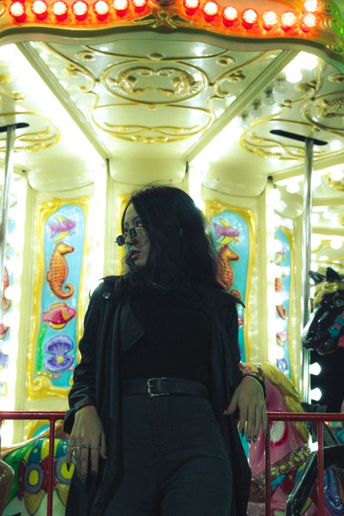 Free A fashionable woman in black attire poses confidently beside a glowing carousel at night. Stock Photo