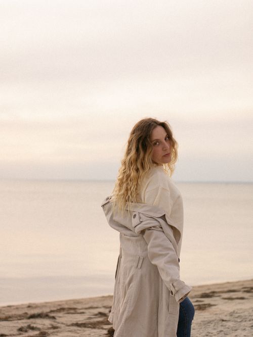 Free A woman in a trench coat gazes at the sunset over a calm seaside view. Stock Photo