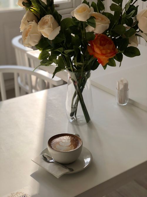 Free A serene café scene featuring a cappuccino and a vase of roses on a sunlit table. Stock Photo