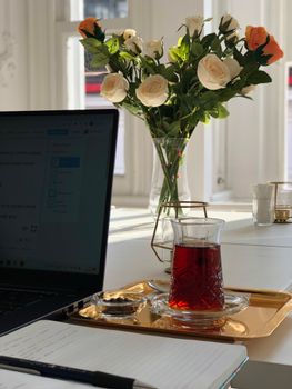 Elegant workspace featuring a glass of tea, notebook, and rose bouquet on a sunlit table.