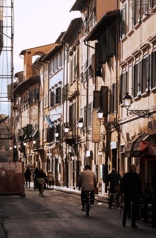 Free Sunlit narrow street in Pisa, Italy, showcasing historic architecture, scooters, and pedestrians. Stock Photo