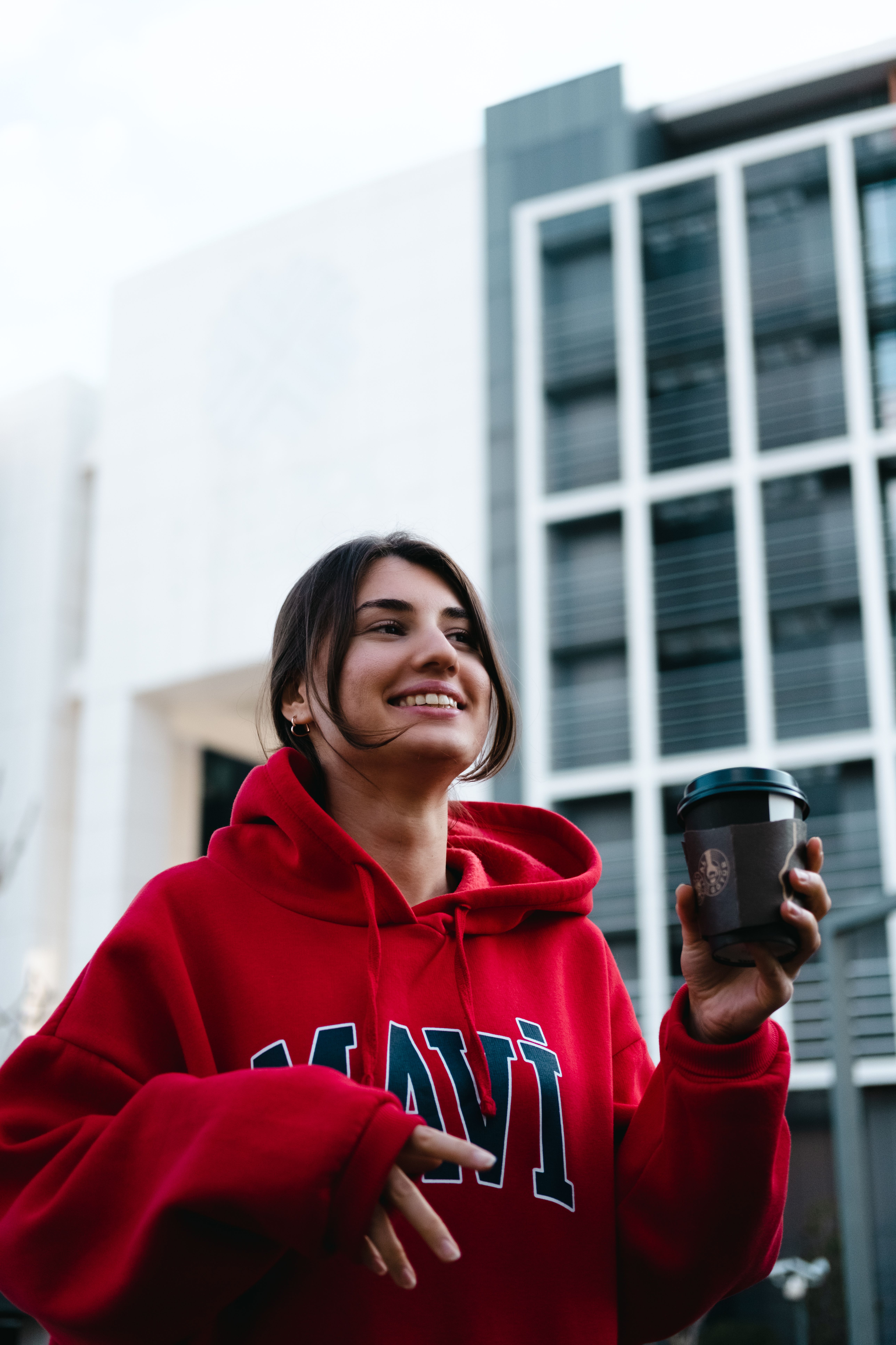 Smiling young woman in a red hoodie holding coffee on a city street.