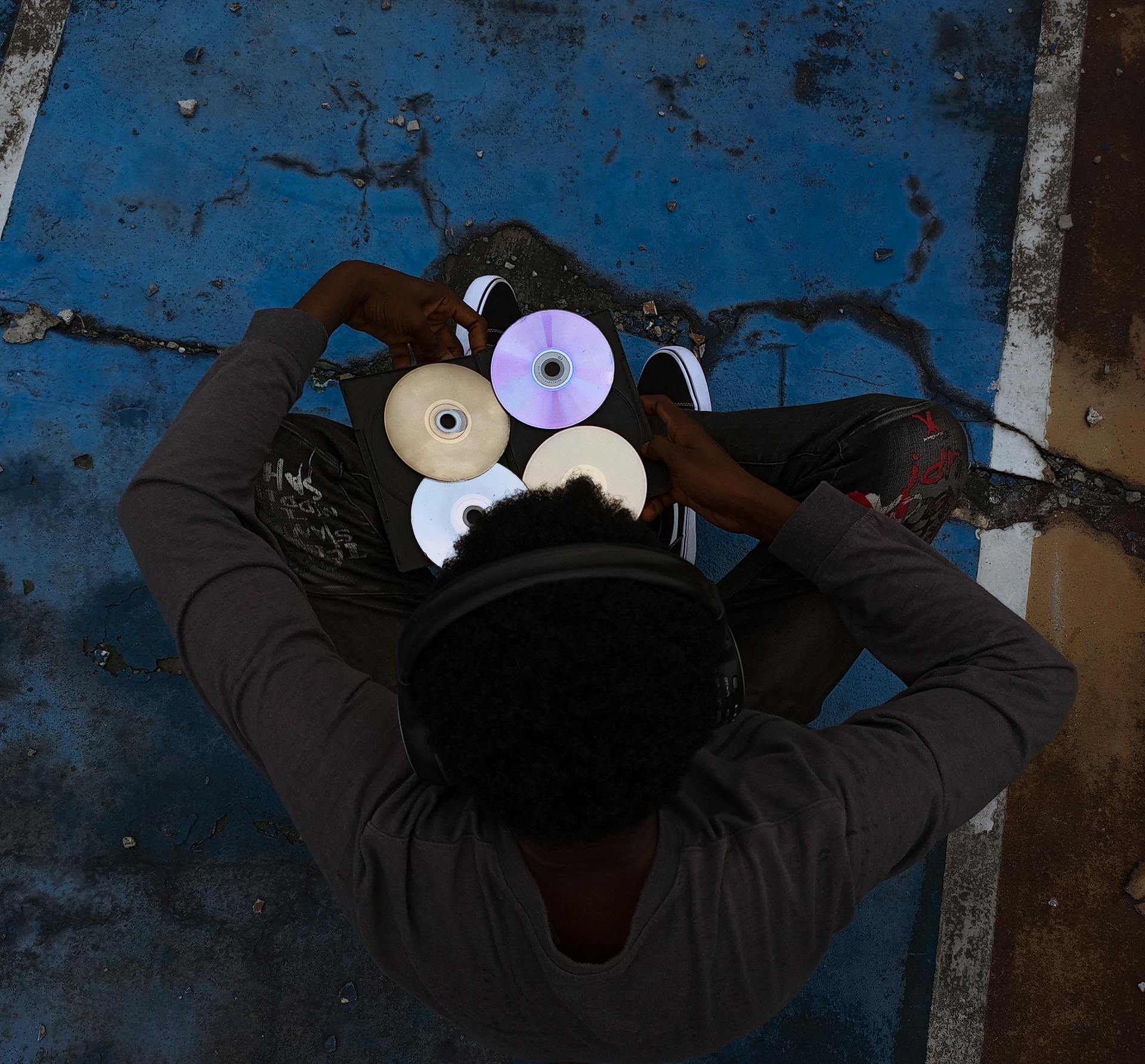 A person in headphones holds CDs while sitting on a cracked blue surface.