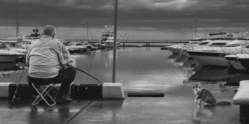 Free A man fishing at Sochi marina with yachts and a cat nearby, captured in black and white. Stock Photo