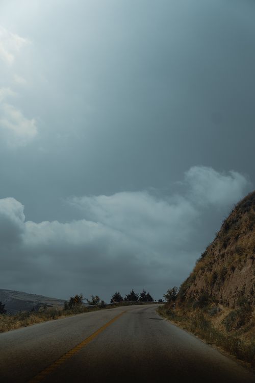 Free A winding rural road stretches into the distance beneath an expansive cloudy sky, bordered by a hillside. Stock Photo