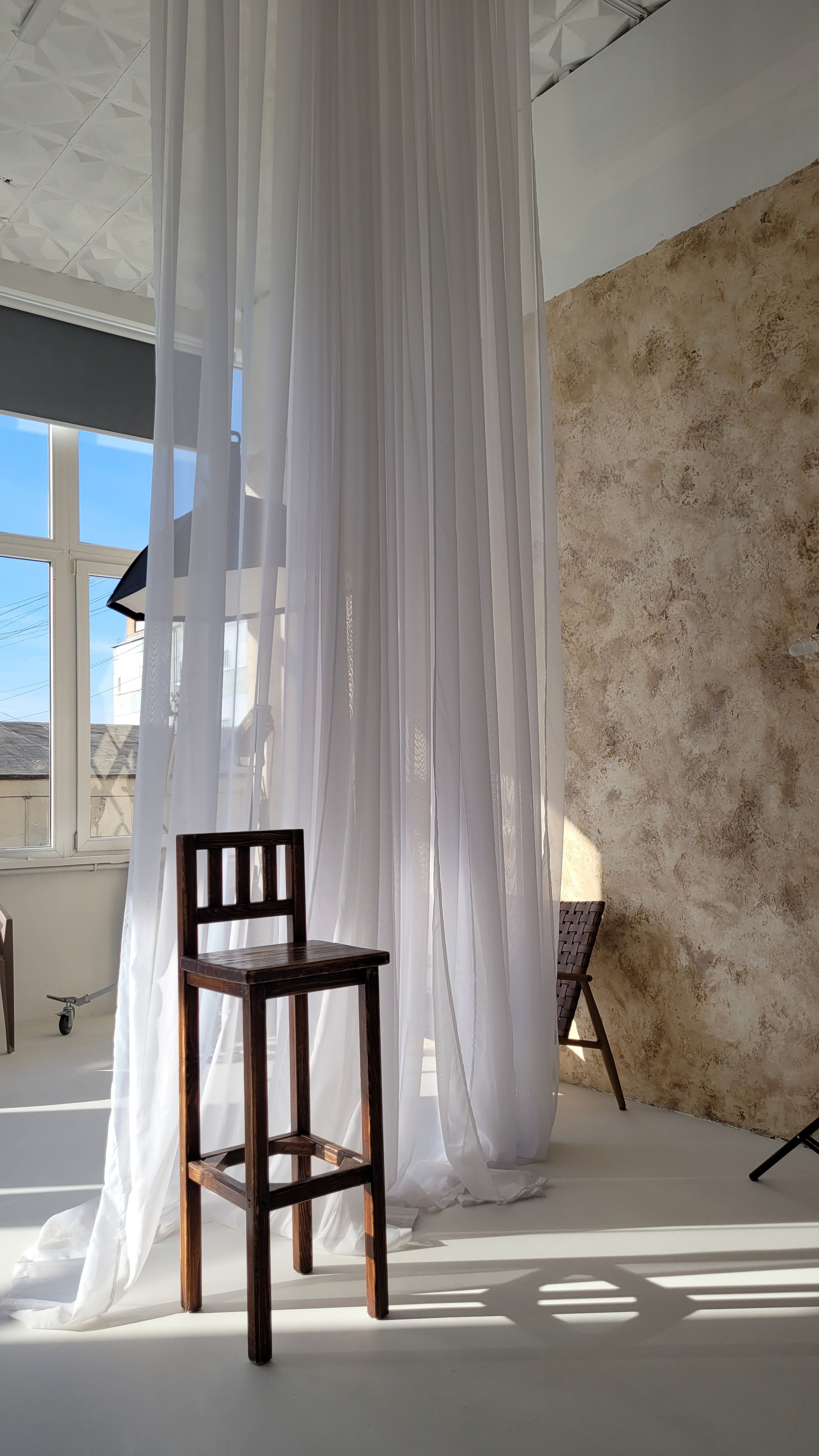 A serene indoor studio setting with a wooden chair and flowing white curtains lit by sunlight.