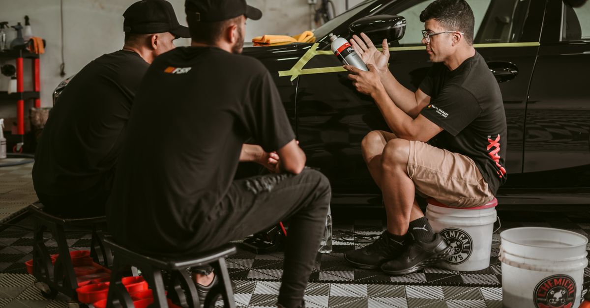 Men discussing car detailing techniques in a professional workshop setting.