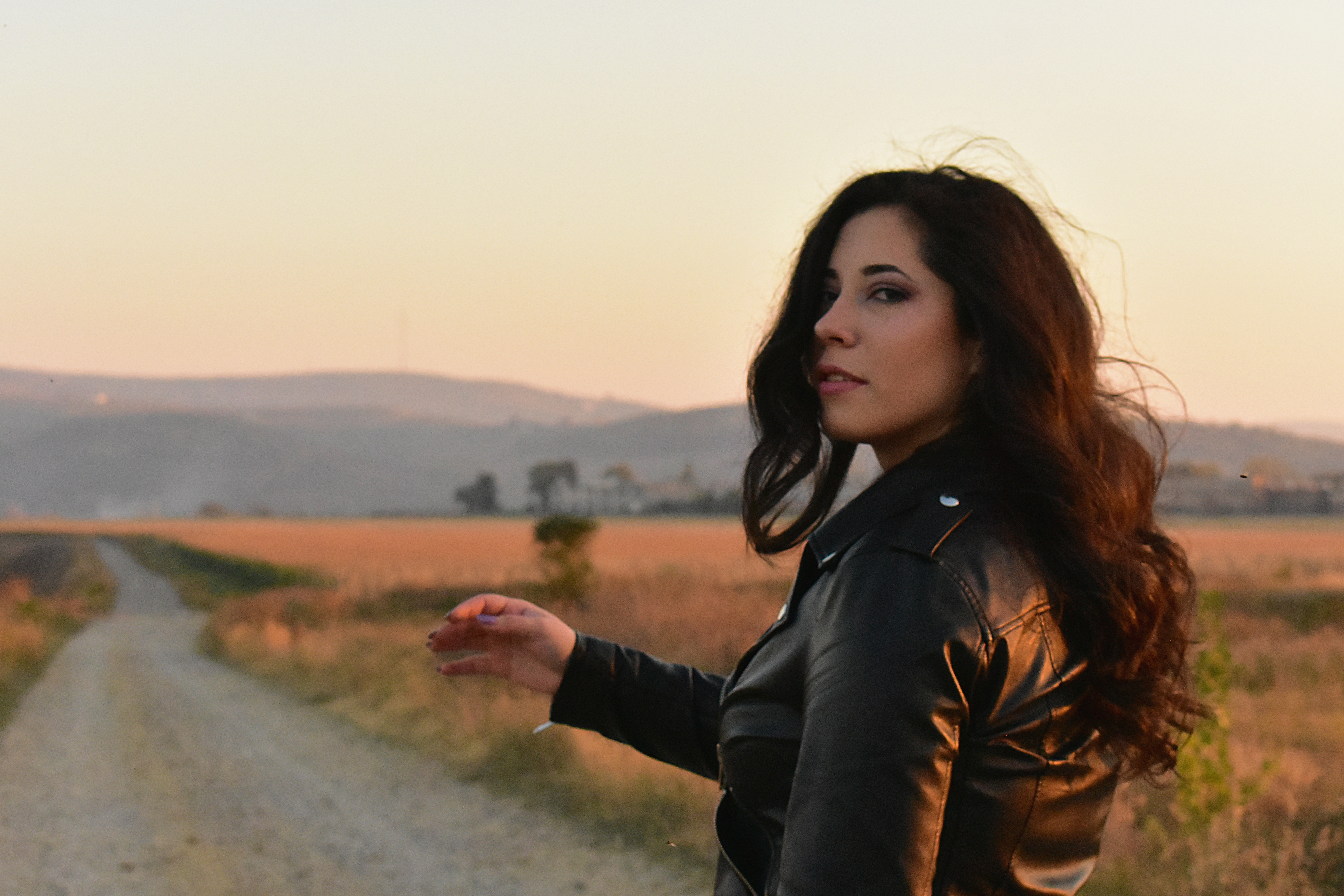 A woman in a black leather jacket stands in a field at sunset, exuding confidence and style.