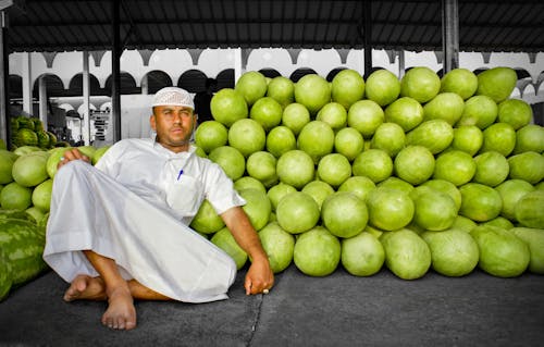 Man in a Gown Sitting next to a Pile of Green Fruit 