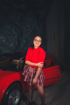 Young woman in a red outfit beside a red sports car, smiling confidently.