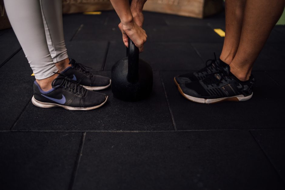 Close-up of two people lifting a kettlebell in a gym setting, emphasizing footwear and strength.