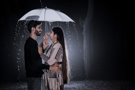A romantic couple smiling and holding an umbrella in the rain at night.