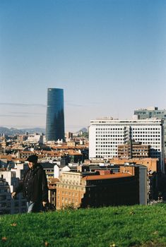 A person in black jacket walks through a grassy area with Bilbao's skyline and iconic tower in the background.