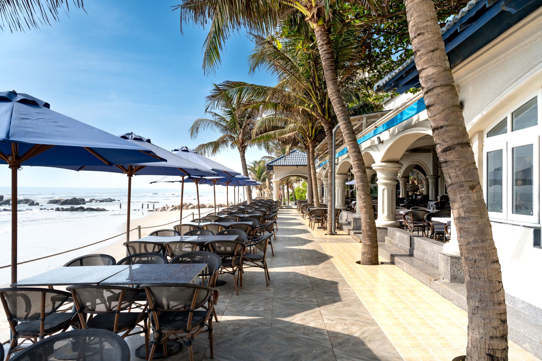 Scenic view of a beachfront patio with tables, umbrellas, and palm trees.