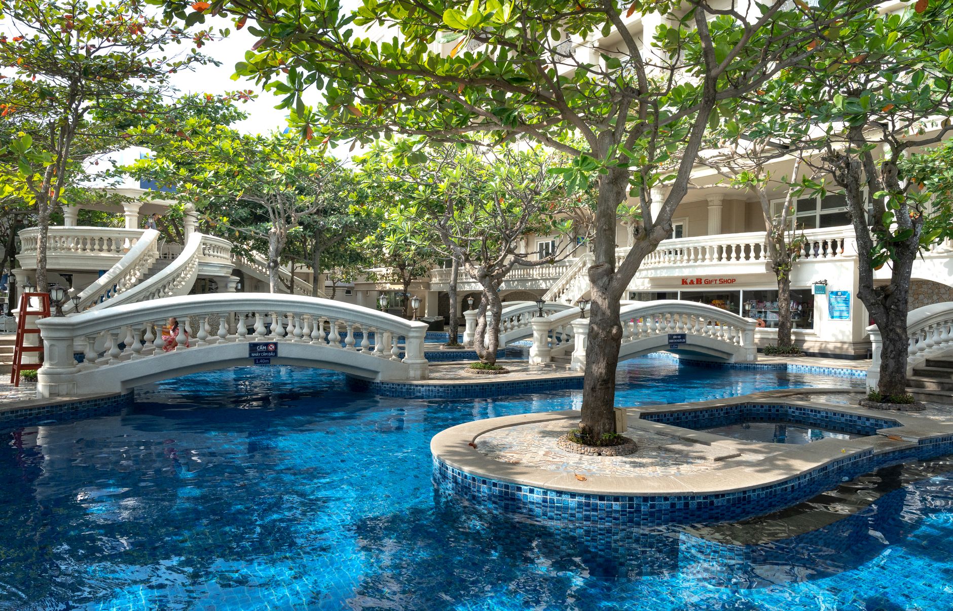 Luxurious holiday resort featuring elegant bridges over a sparkling blue swimming pool surrounded by trees.