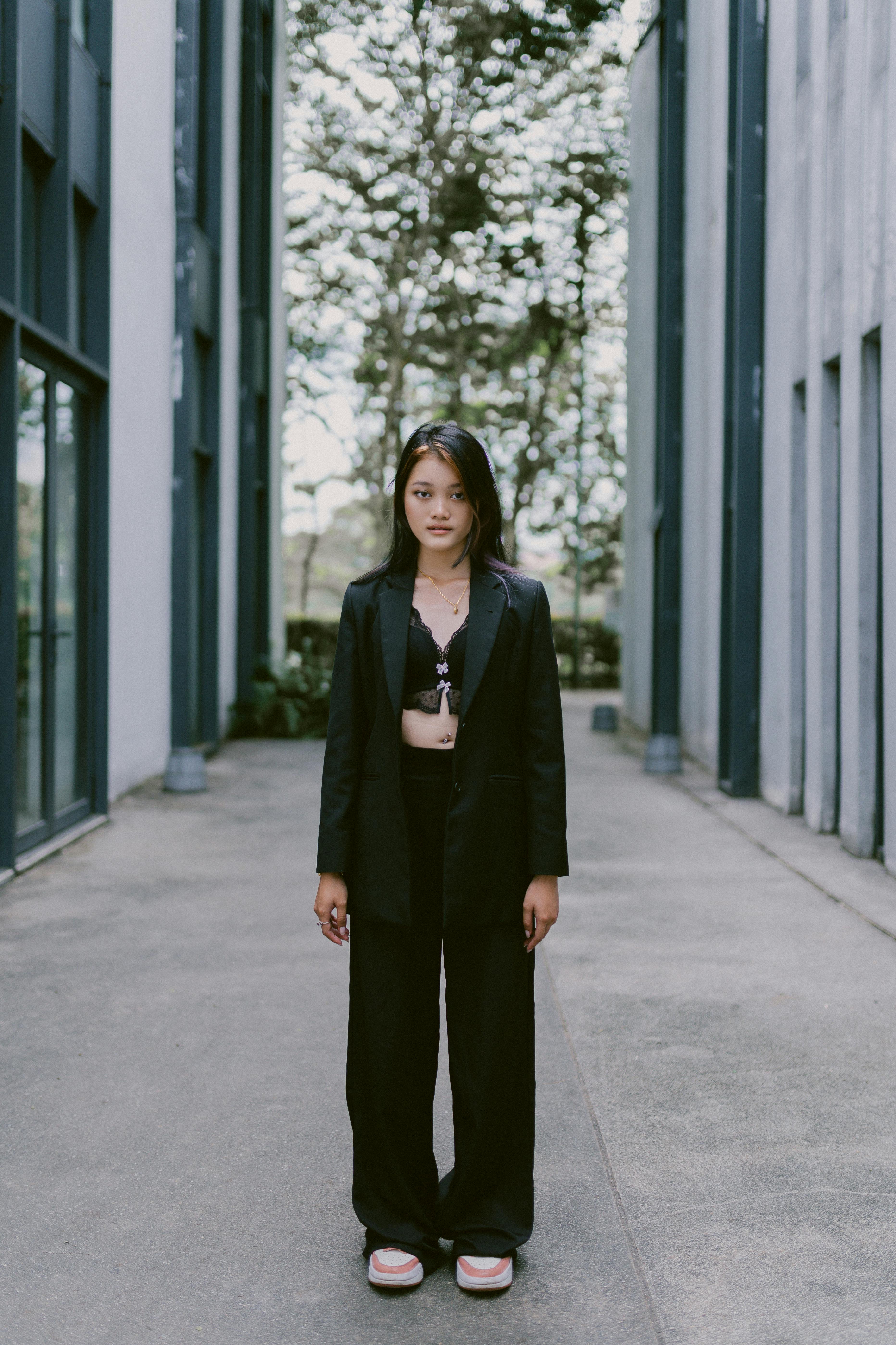 Asian woman in black blazer and pants standing outdoors, showcasing modern style amidst urban architecture.