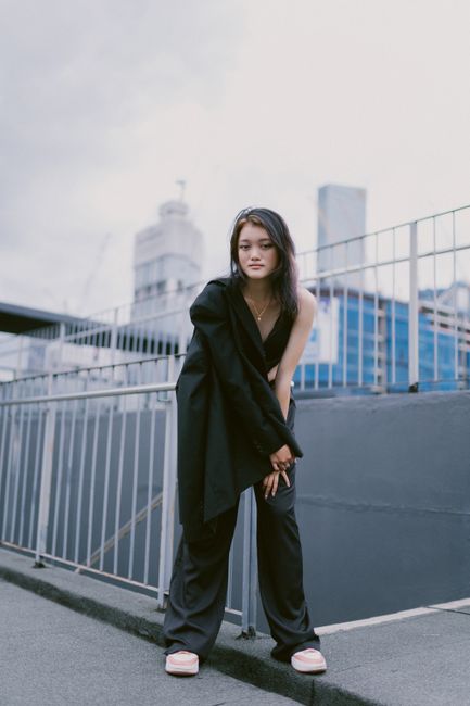 Young Asian woman in fashionable black outfit posing outdoors with city backdrop.