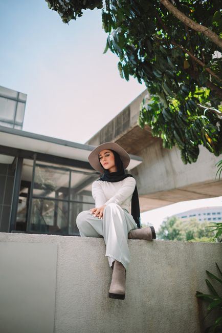 Elegant portrait of a woman in a hat and headscarf, seated against a modern urban backdrop.