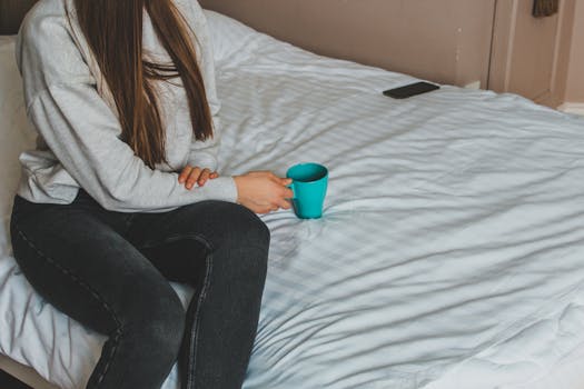 A woman holds a blue mug while sitting on a bed indoors.