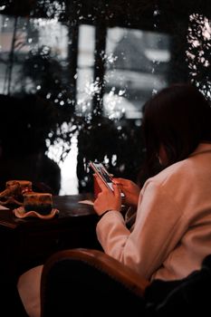 A woman in a cozy cafe uses her smartphone by the window on a rainy day.