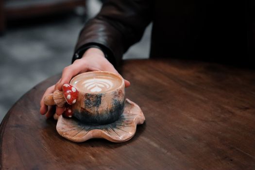 Close-up of a unique ceramic coffee cup with latte art, held by a hand on a wooden table.