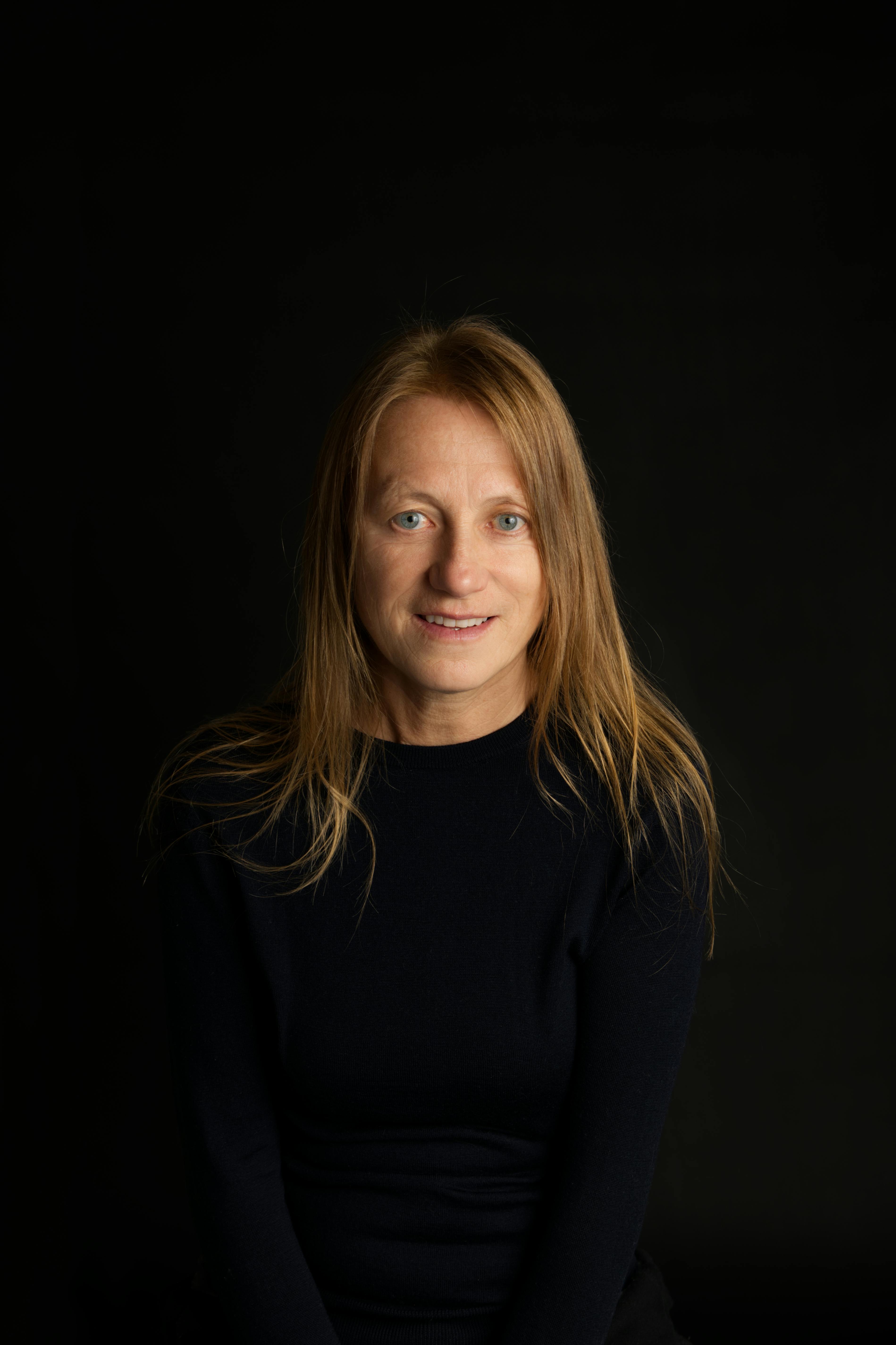 Studio portrait of a woman smiling against a black background, wearing a dark sweater.