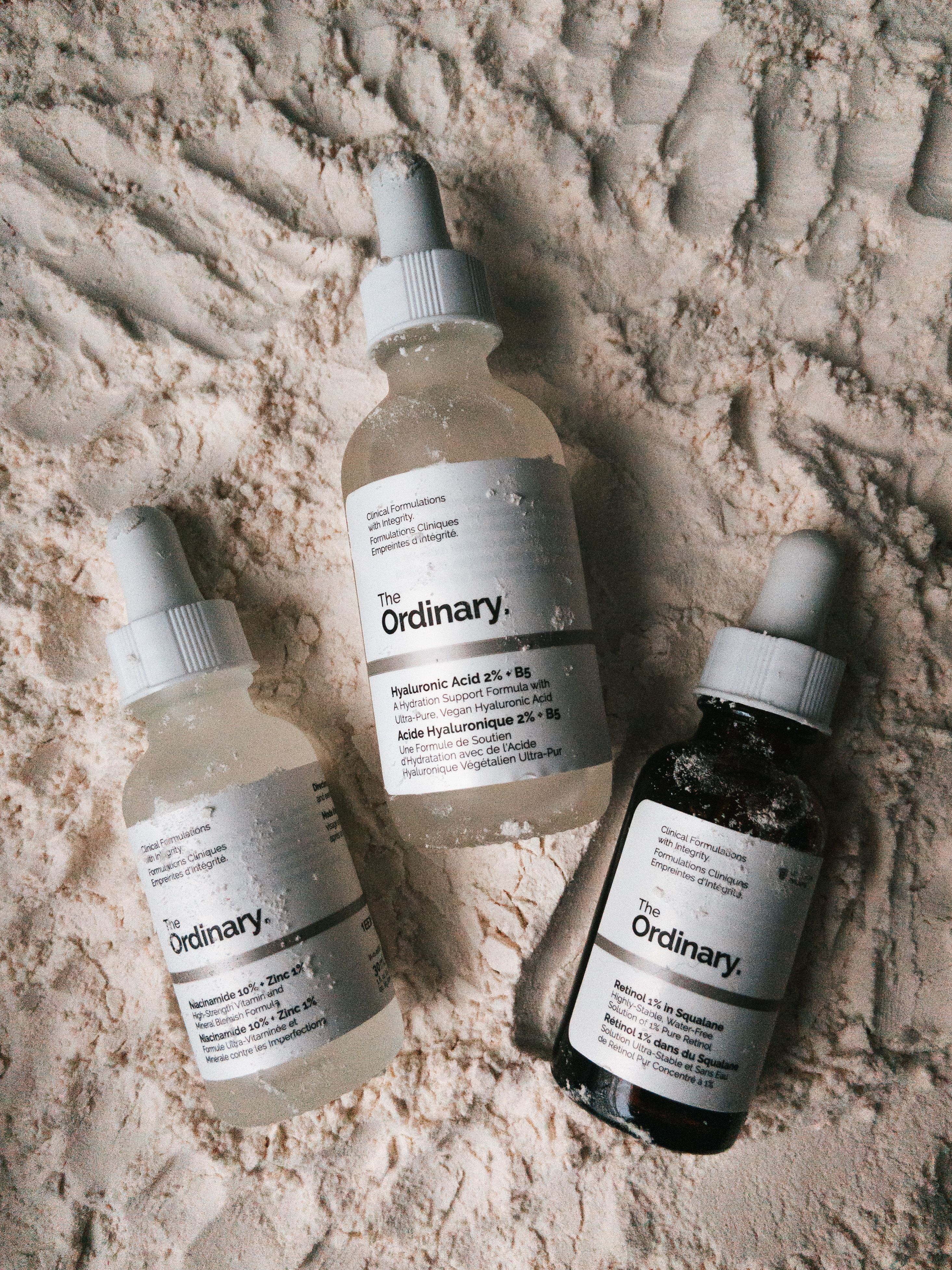 Três frascos conta-gotas de séruns da The Ordinary exibidos sobre uma superfície dos pós.