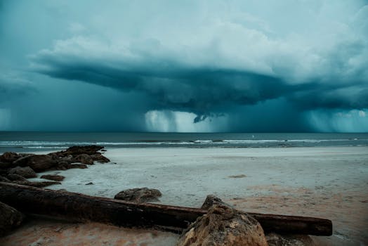 Turbulent storm clouds loom over a quiet beach with driftwood and rocks in view.
