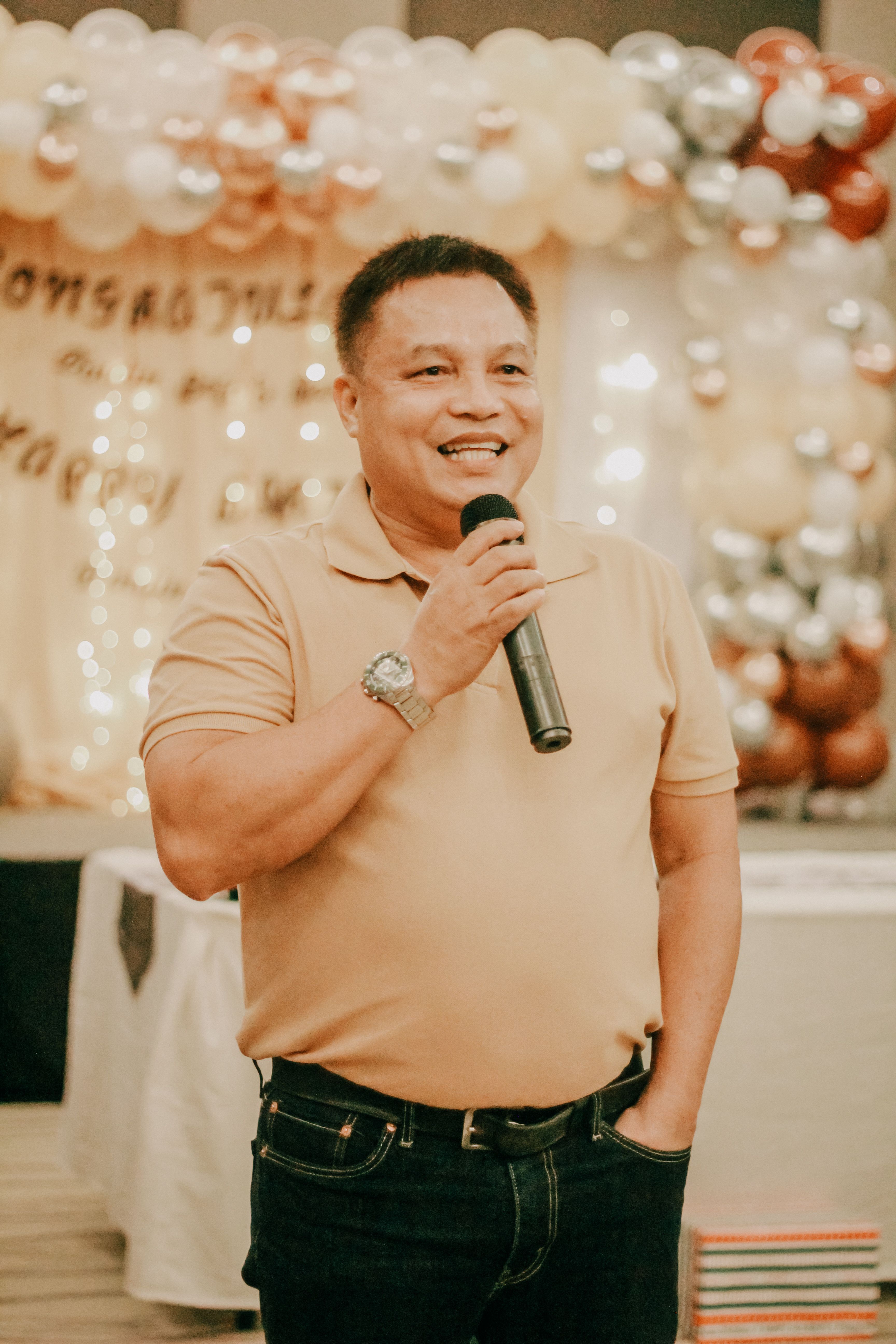 Mid adult Asian man speaking into a microphone at an indoor celebration event with a balloon backdrop.