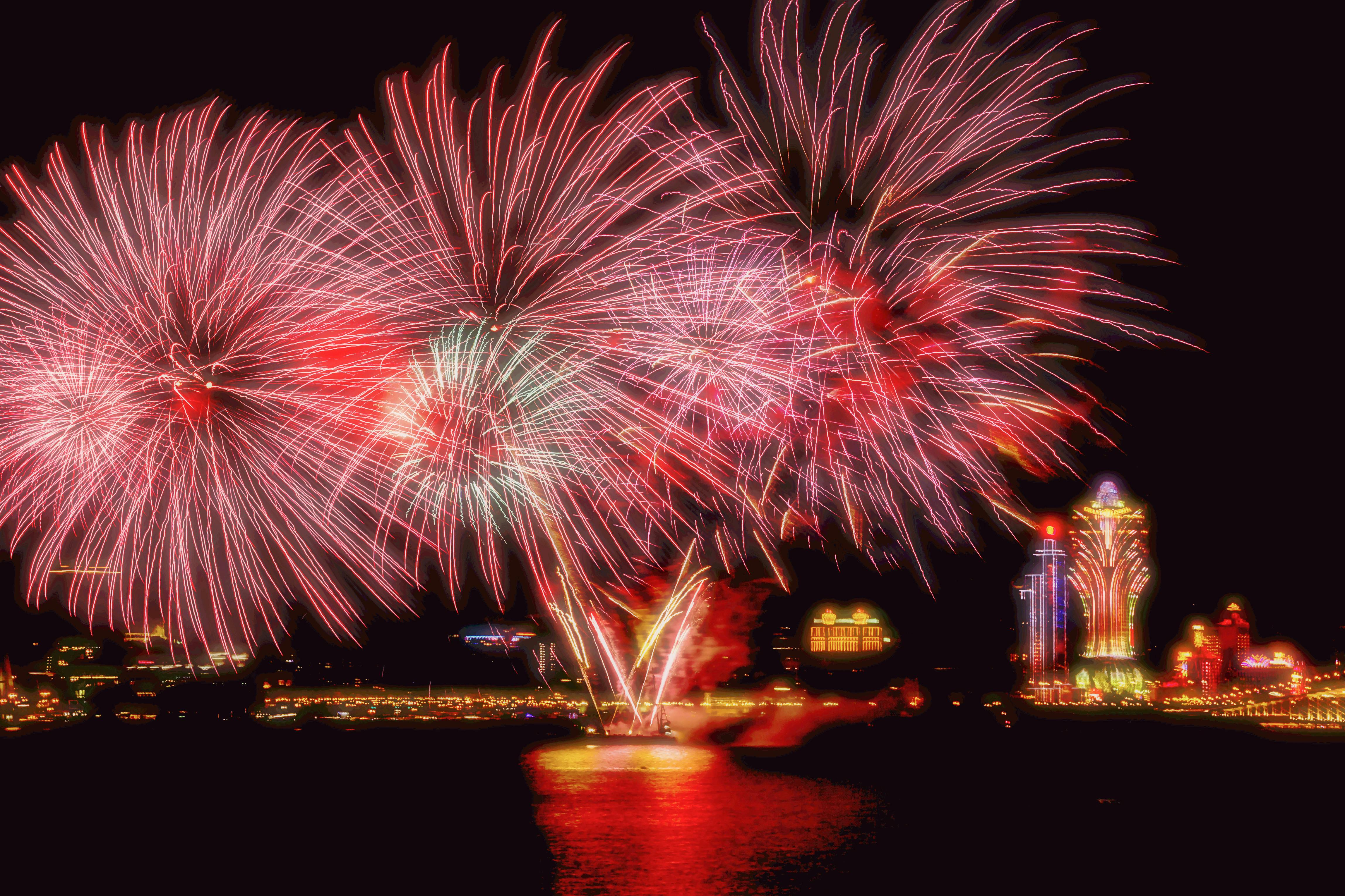 Vibrant pink and red fireworks light up the night sky over Macau's iconic skyline, reflecting on the water.