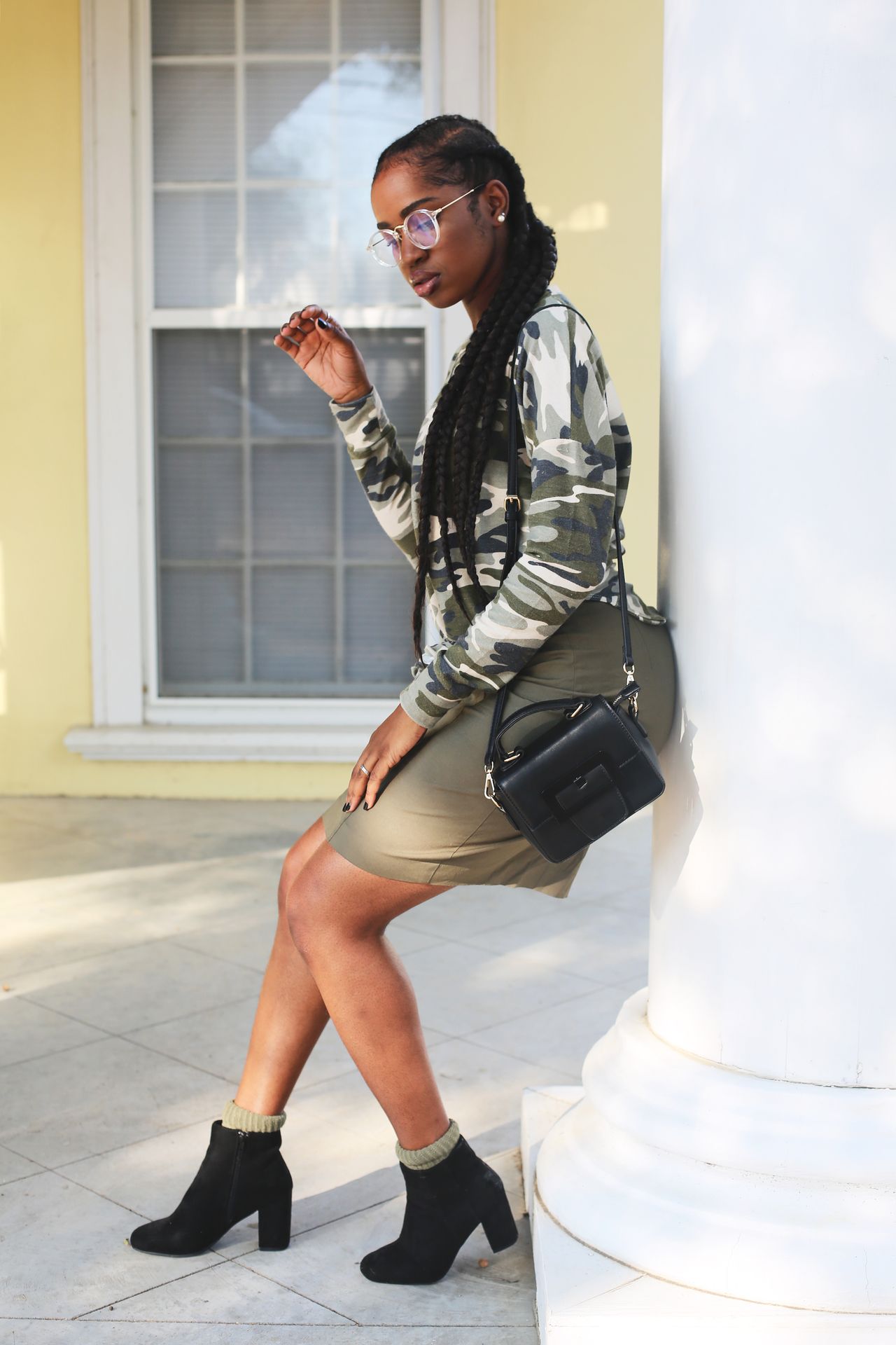 Woman in a camo top, khaki mini skirt and black ankle boots leaning on a pillar