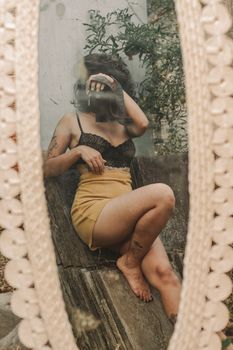 A woman with tattoos takes a self-portrait through a vintage mirror, showcasing style and creativity.