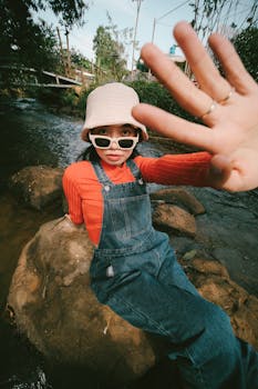 Fashionable woman in denim overalls sitting by a stream, wearing sunglasses and a hat.