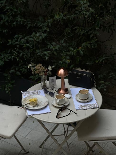 Intimate outdoor coffee setting on a white table with desserts, two cups, and ambient lighting, surrounded by foliage.