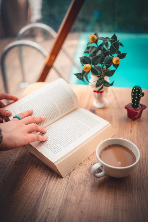 Photo of an Open Book and a Cup of Coffee on a Table by the Pool